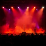 Flick International Empty concert stage with guitar stand and microphone, surrounded by colorful lights and smoke from an electrical fire