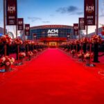 Flick International A vibrant red carpet scene at the 2025 ACM Awards featuring black stanchions and floral arrangements