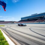 Flick International Race car navigating the Indianapolis Motor Speedway oval track with racing flags in the foreground