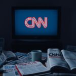 Flick International A dimly lit room with a muted television displaying the CNN logo, surrounded by newspapers about President Biden's mental decline and cancer diagnosis.