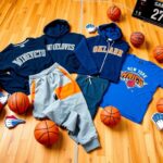 Flick International Collection of NBA team merchandise including Timberwolves sweatshirt, Thunder jogger sweatpants, Knicks hoodie, and Pacers t-shirt on a wooden surface