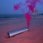 Flick International Silver military flare partially buried in the sand at a Lake Michigan beach with gentle red smoke.