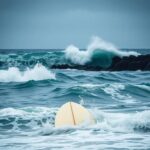 Flick International Tumultuous ocean waves crashing against rocks with a surfboard in the foreground