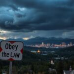Flick International Dramatic landscape of Colorado featuring a weathered road sign that reads 'Obey the Law' against the backdrop of the Rocky Mountains and Denver's skyline.