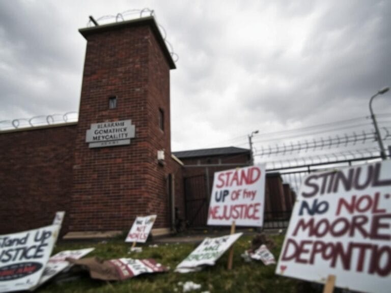 Flick International A dramatic scene outside a Newark immigration facility showing a strong brick wall with barbed wire and protest signs.